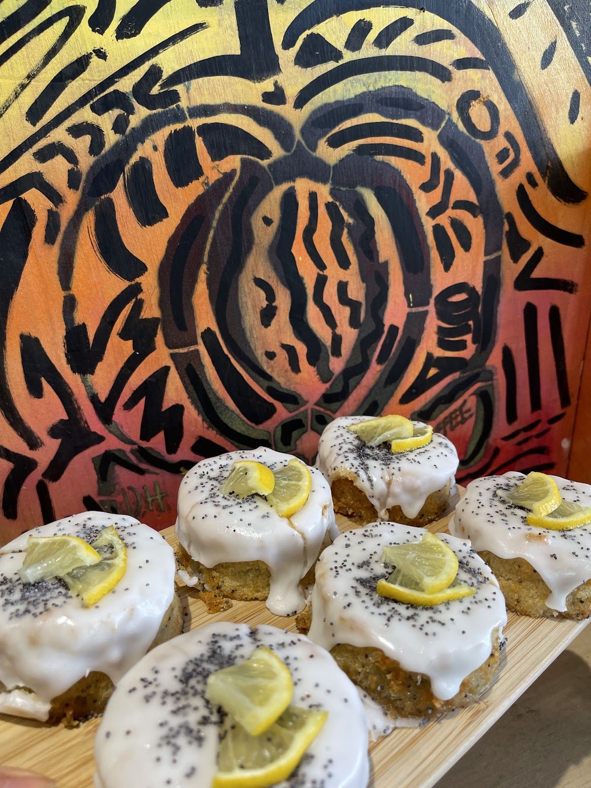 Round cakes with white icing, poppy seeds and lemon slices on a board in front of the orange and black painted mural.