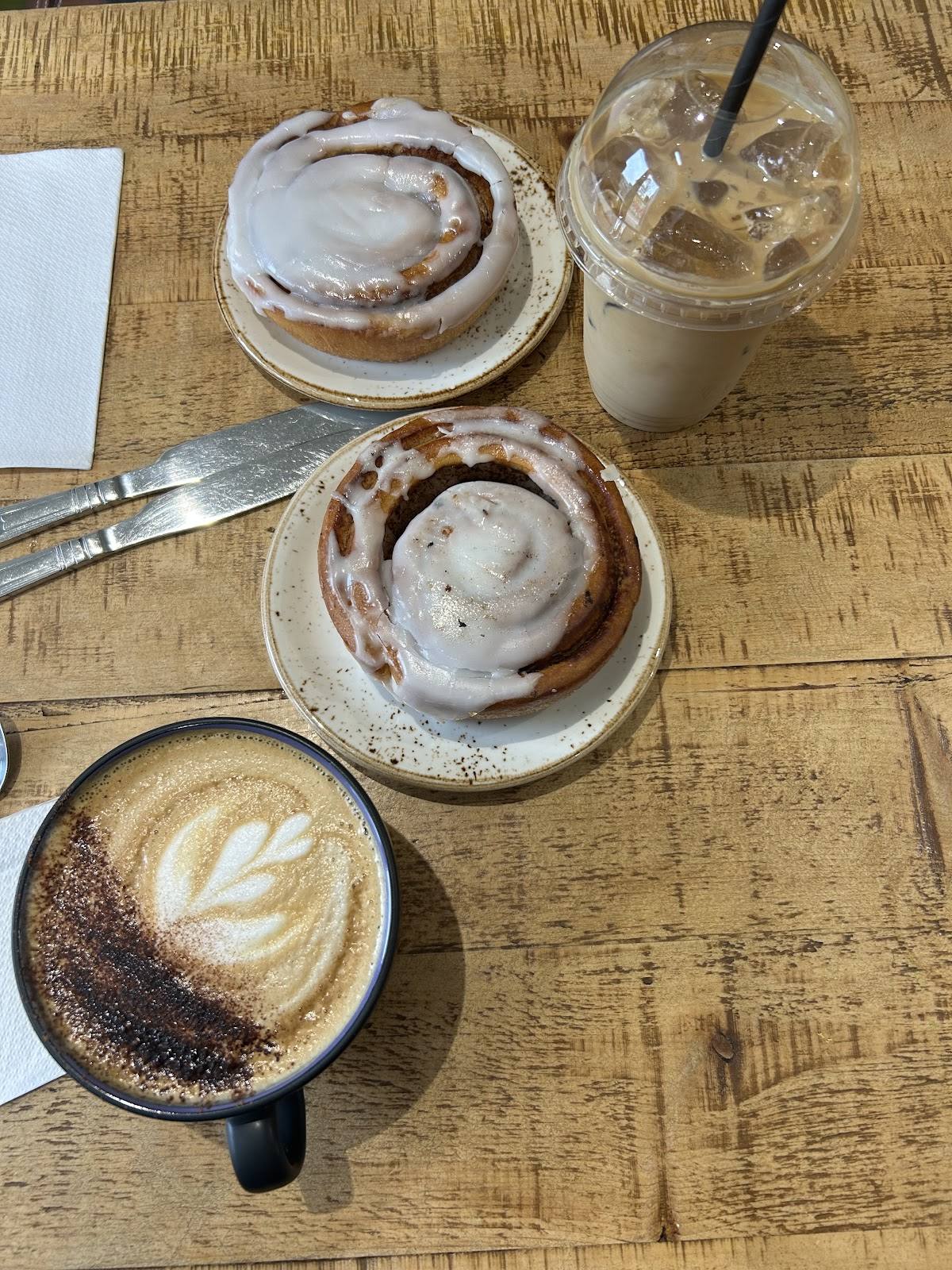 Two iced cinnamon buns, an iced coffee and a latte with a rosetta on a rustic wooden table.