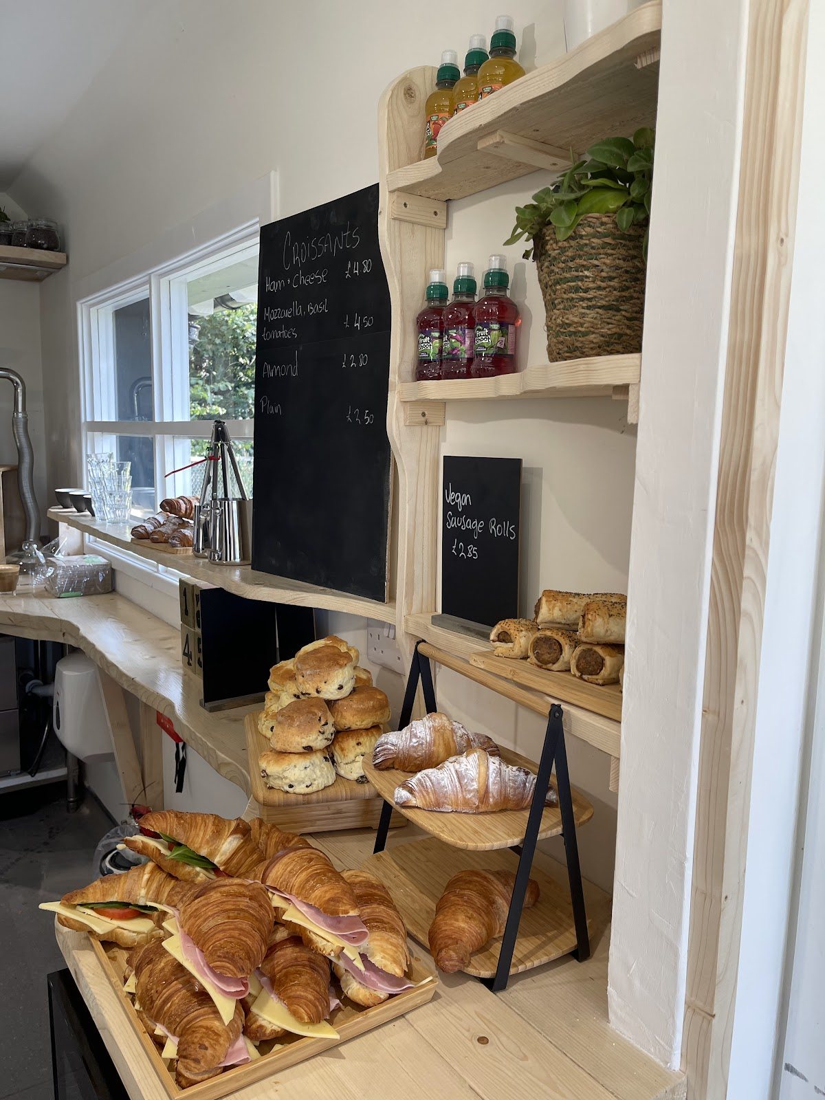 Pale pine shelves with chalkboards for croissants and vegan sausage rolls, above trays of fruit scones, croissants, filled croissants and seeded sausage rolls.
