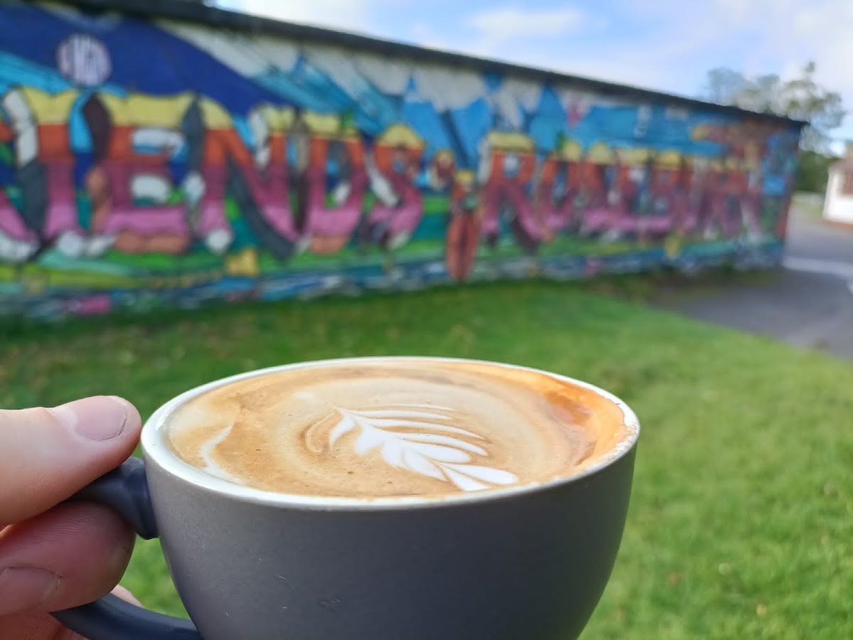 A flat white with latte art in a grey cup, held up against the grass and a colourful graffiti mural reading 'Friends of Roseburn…' in the park.